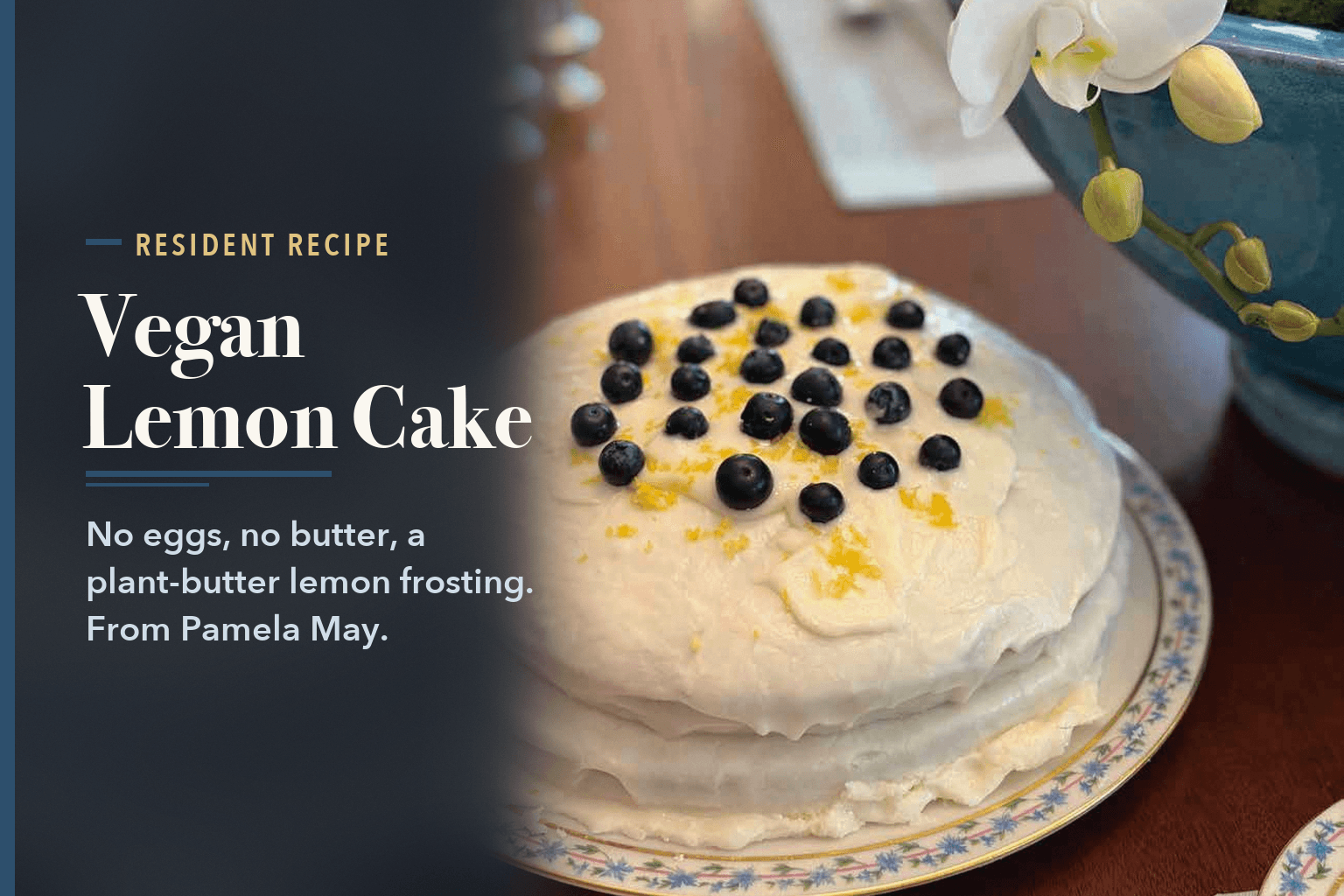 A frosted two-layer lemon cake topped with blueberries and lemon zest on a floral-rimmed china plate, a white orchid behind it on a wooden table. No people in it, beside the words Vegan Lemon Cake With Lemon Buttercream Frosting.
